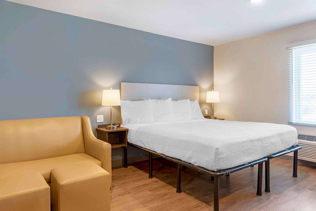 A hotel room with a bed, two bedside lamps, a sofa, and a window with blinds.