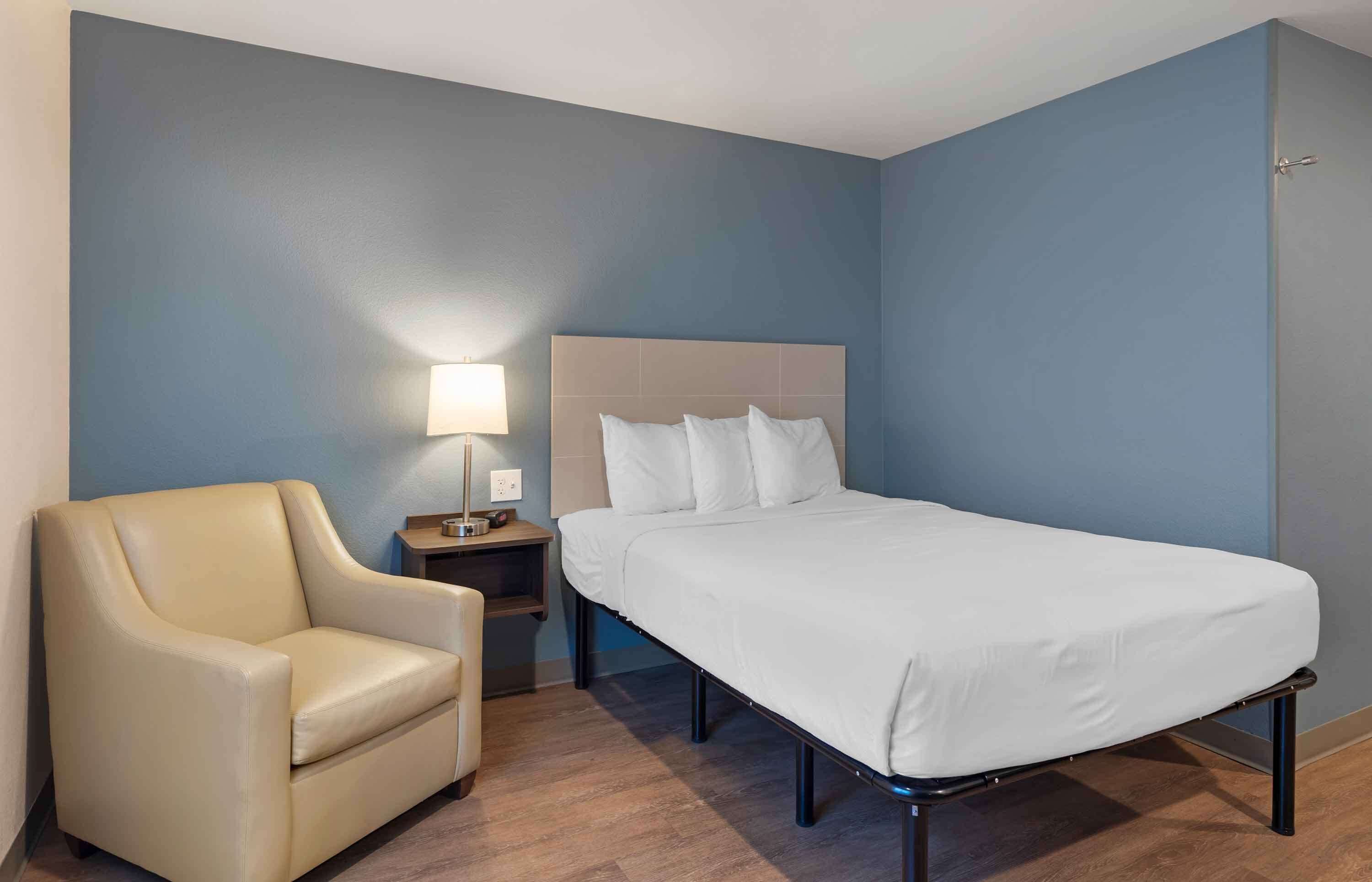 Una habitación de hotel con una cama, una silla, una mesita de noche y una lámpara.
