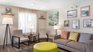 Una sala de estar moderna con un sofá, dos sillones, una mesa de centro y una lámpara de pie. Se observan cuadros enmarcados en las paredes y una ventana con cortinas.