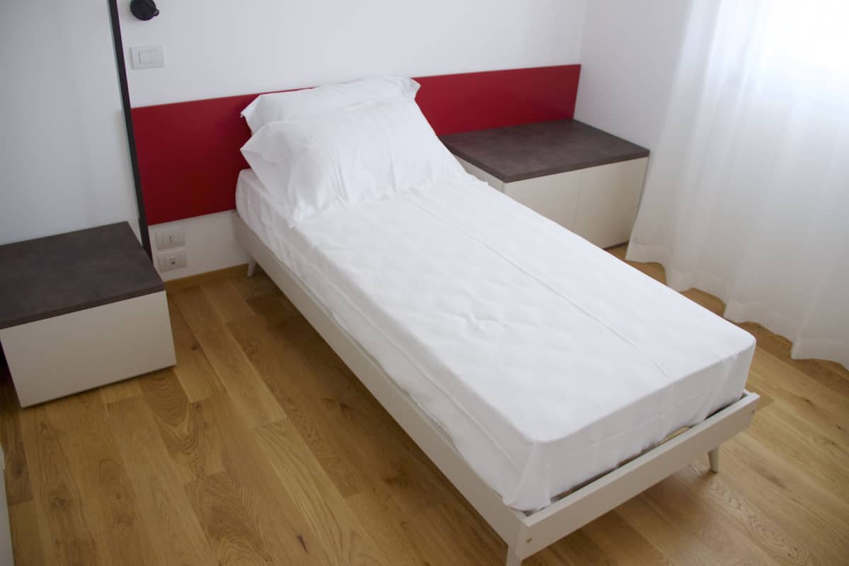 A single bed with a white mattress and red headboard in a room with wooden flooring and a small bedside table.