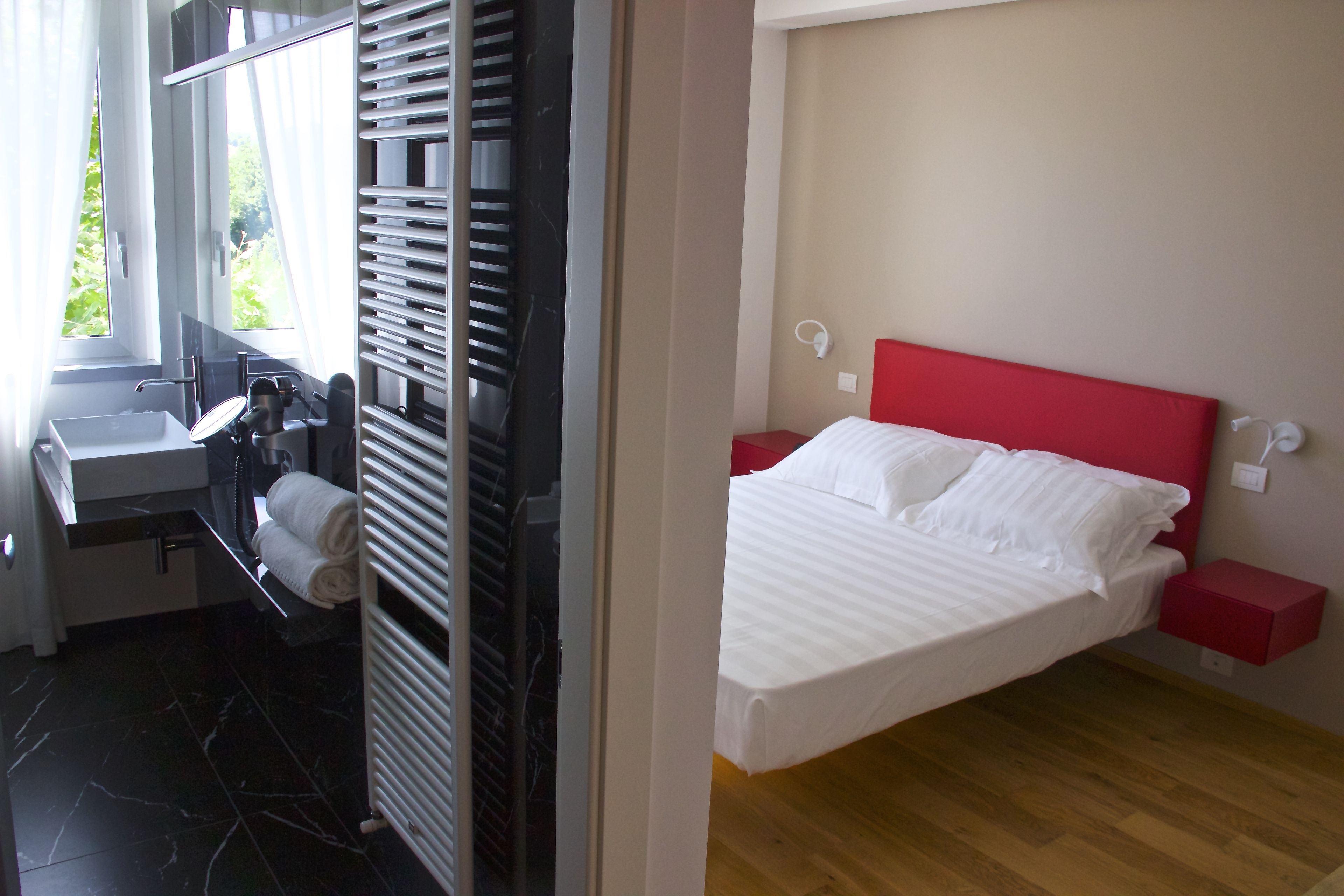 Ein Hotelzimmer mit Bett, rotem Kopfteil, Nachttisch, rotem Beistelltisch, weißer Nachttischlampe, Fenster mit Vorhängen, Badezimmer mit Waschbecken, Spiegel, Handtuchhalter und Heizkörper.