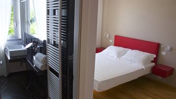 Ein Hotelzimmer mit Bett, rotem Kopfteil, Nachttisch, rotem Beistelltisch, weißer Nachttischlampe, Fenster mit Vorhängen, Badezimmer mit Waschbecken, Spiegel, Handtuchhalter und Heizkörper.