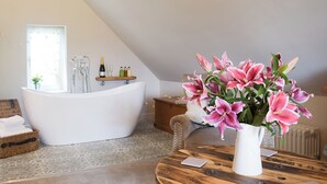 Une salle de bain avec une baignoire sur pieds, une étagère en bois avec des articles de toilette, un vase avec des fleurs et un radiateur.