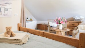 Une chambre à coucher avec un lit, une tête de lit en bois, une table de chevet ornée d’un vase de fleurs, une chaise, un canapé, une commode en bois et une baignoire.