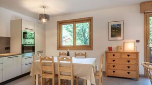 A modern kitchen with a dining table and chairs, a wooden dresser, and a window with a view of trees.