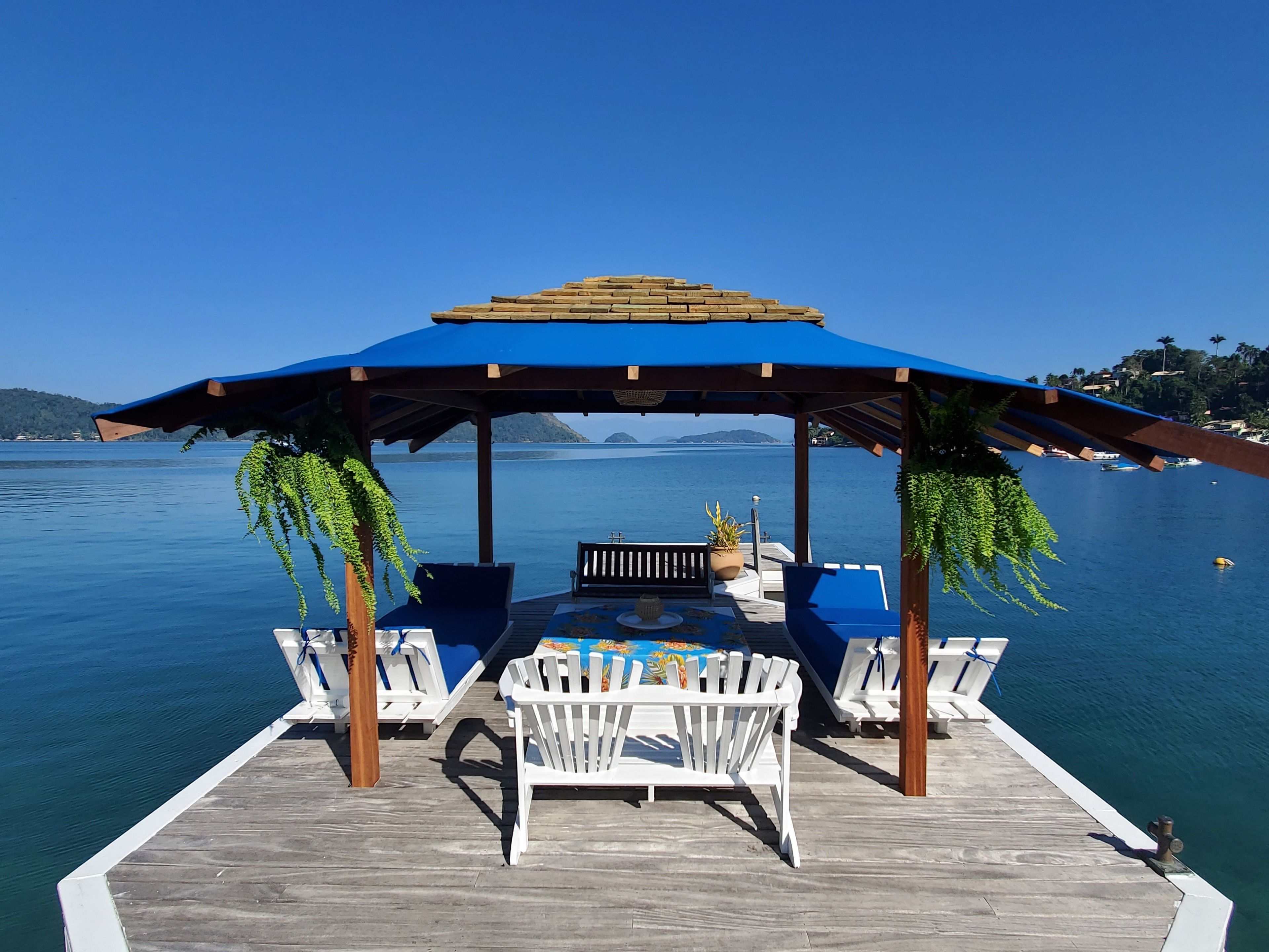 Un muelle de madera con un toldo azul, sillas de lounge blancas y una mesa con mantel de colores.