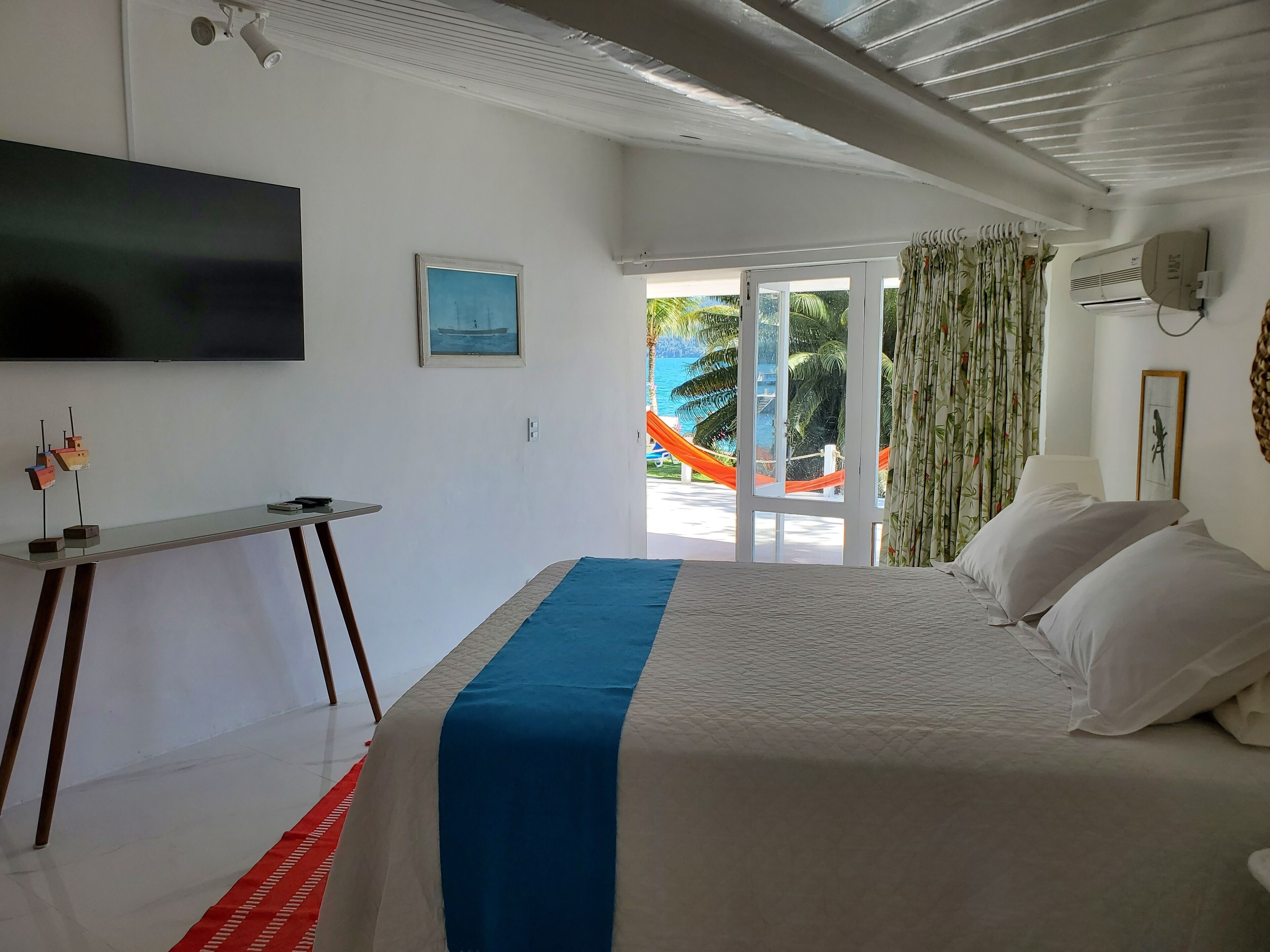 Ein Hotelzimmer mit Bett, Schreibtisch, Fernseher und Meerblick durch die Tür.