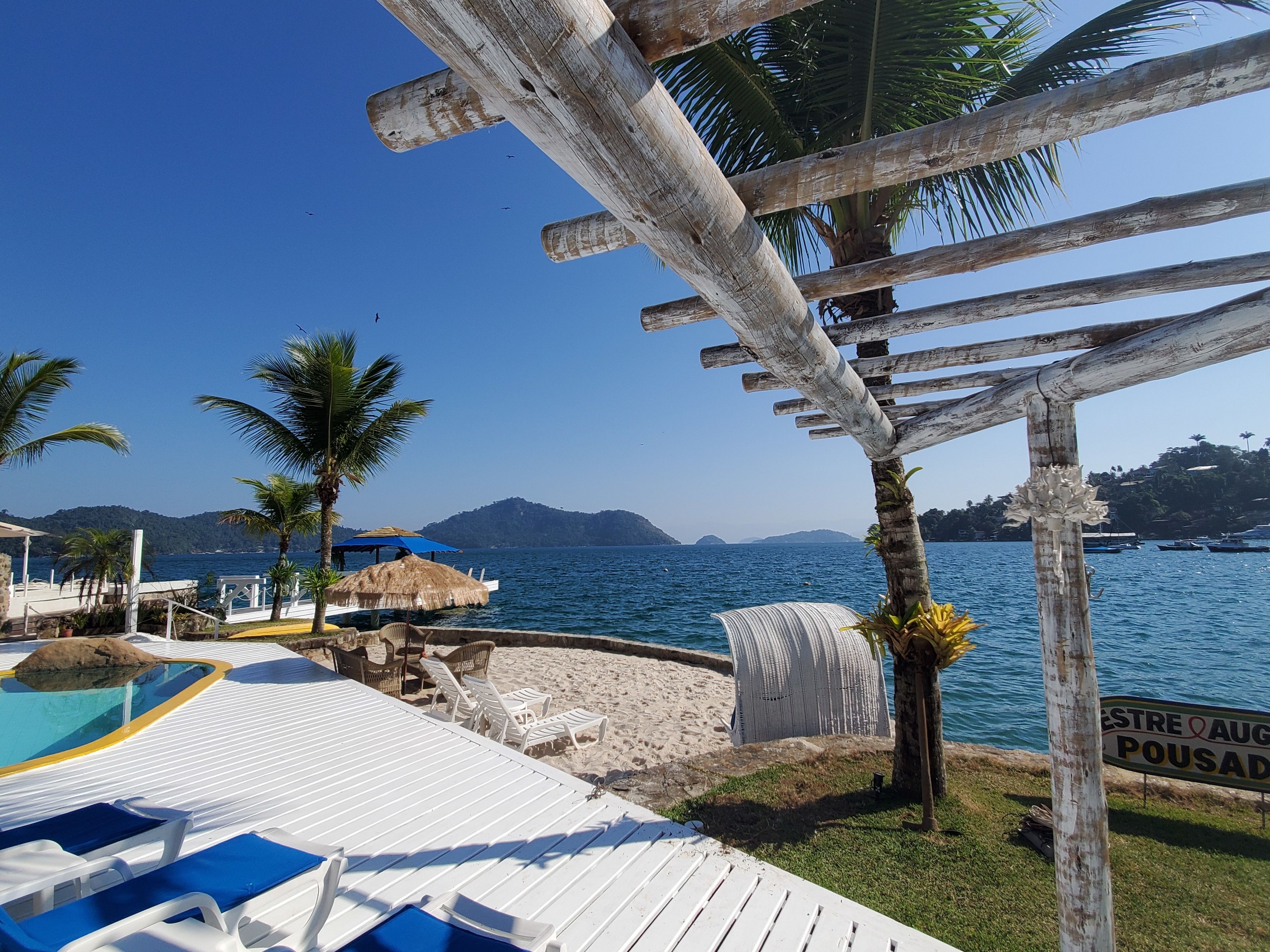 Uma área de piscina à beira-mar com espreguiçadeiras, um guarda-sol de palha e uma pérgola de madeira.