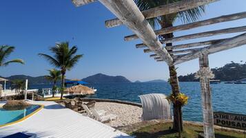 Uma área de piscina à beira-mar com espreguiçadeiras, um guarda-sol de palha e uma pérgola de madeira.