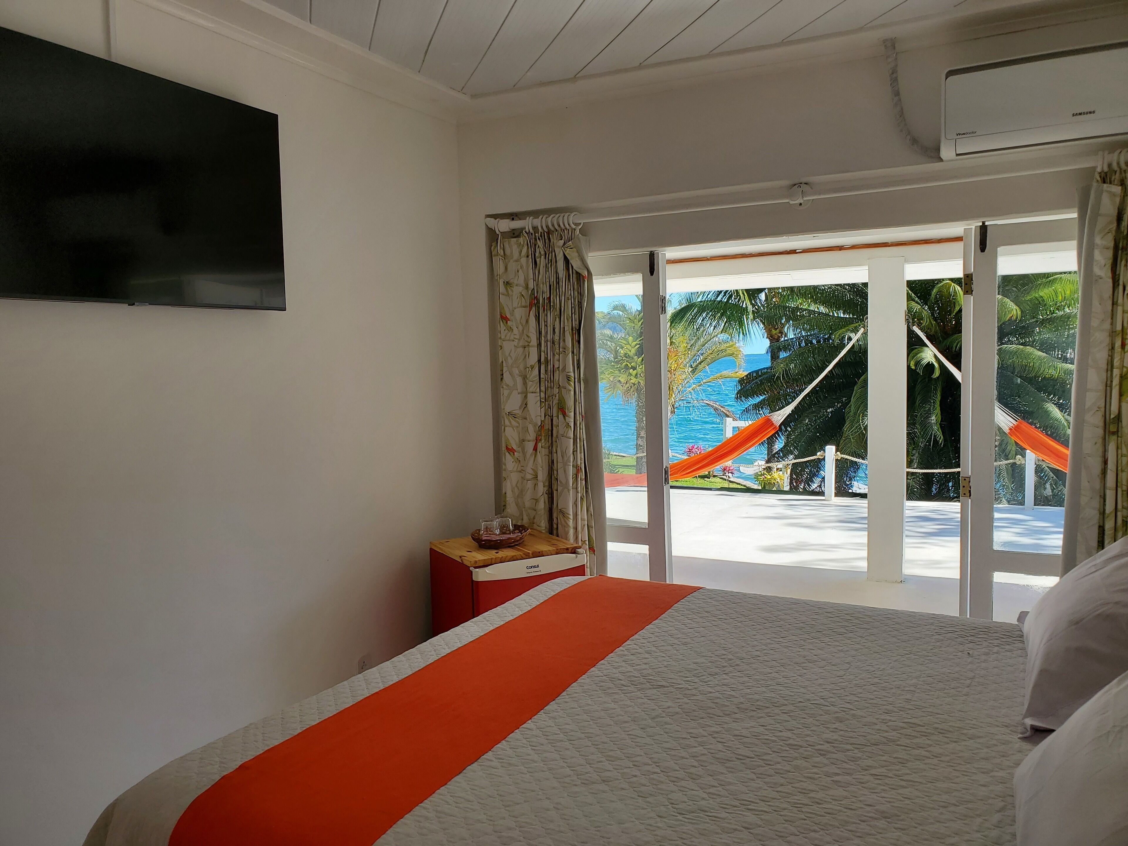Quarto de hotel com cama, televisão na parede e vista para a área da piscina com uma rede.