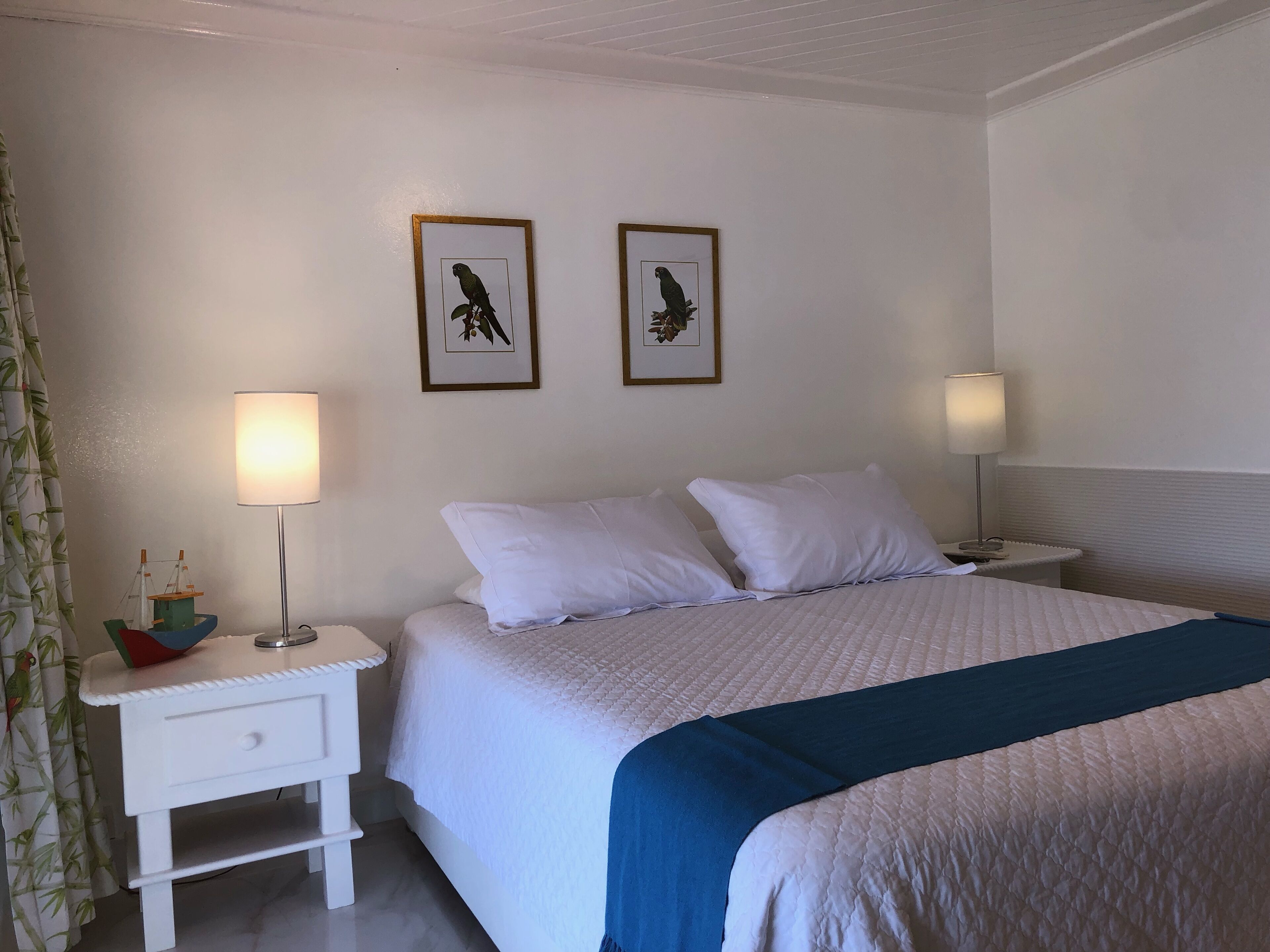 Uma cama bem arrumada com roupa de cama branca e um toque de azul, ladeada por mesas de cabeceira brancas com luminárias, e duas ilustrações de pássaros emolduradas na parede.