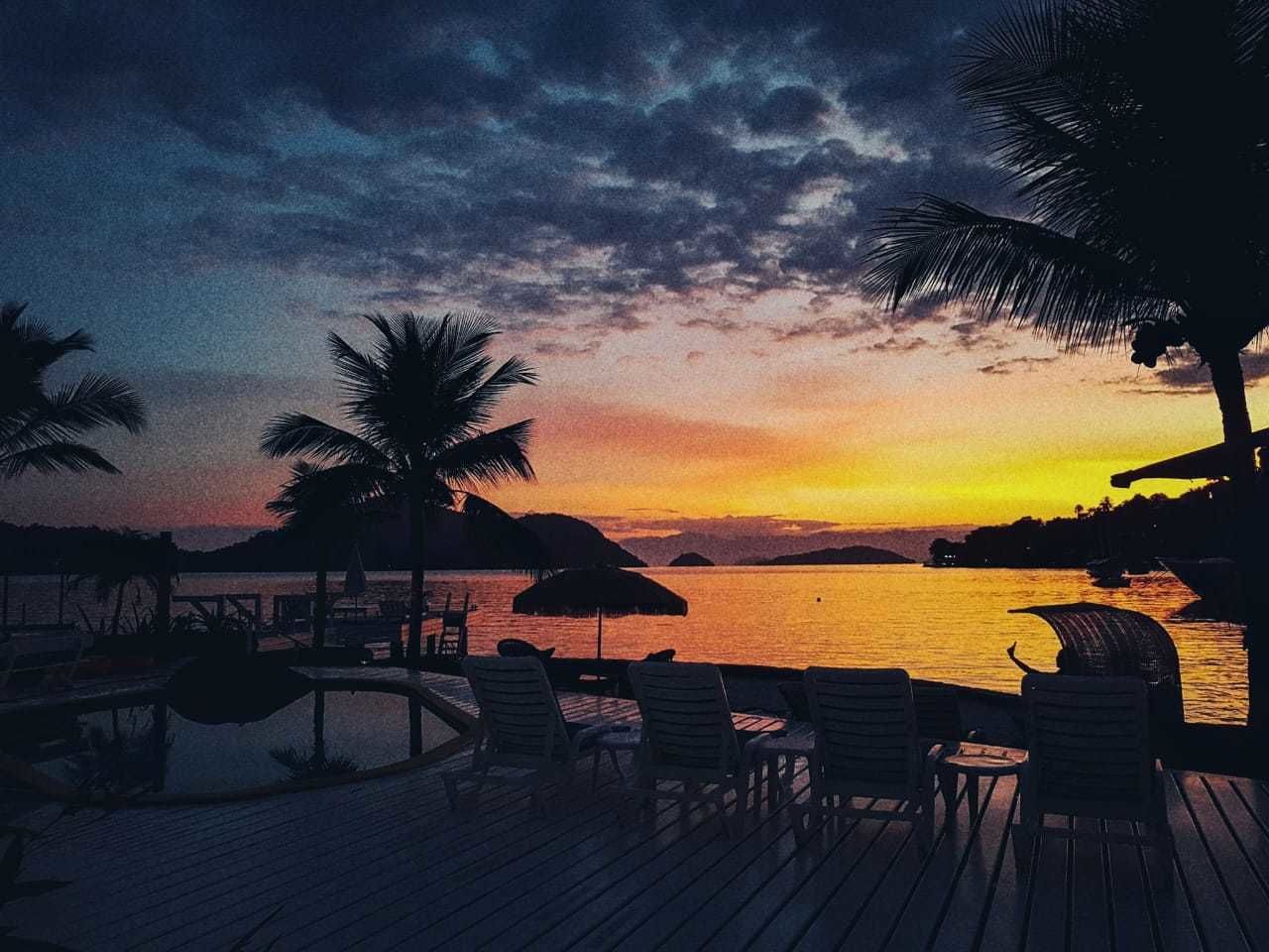 Vista del atardecer desde una terraza con sillas de descanso y sombrillas, con vista a una masa de agua tranquila.