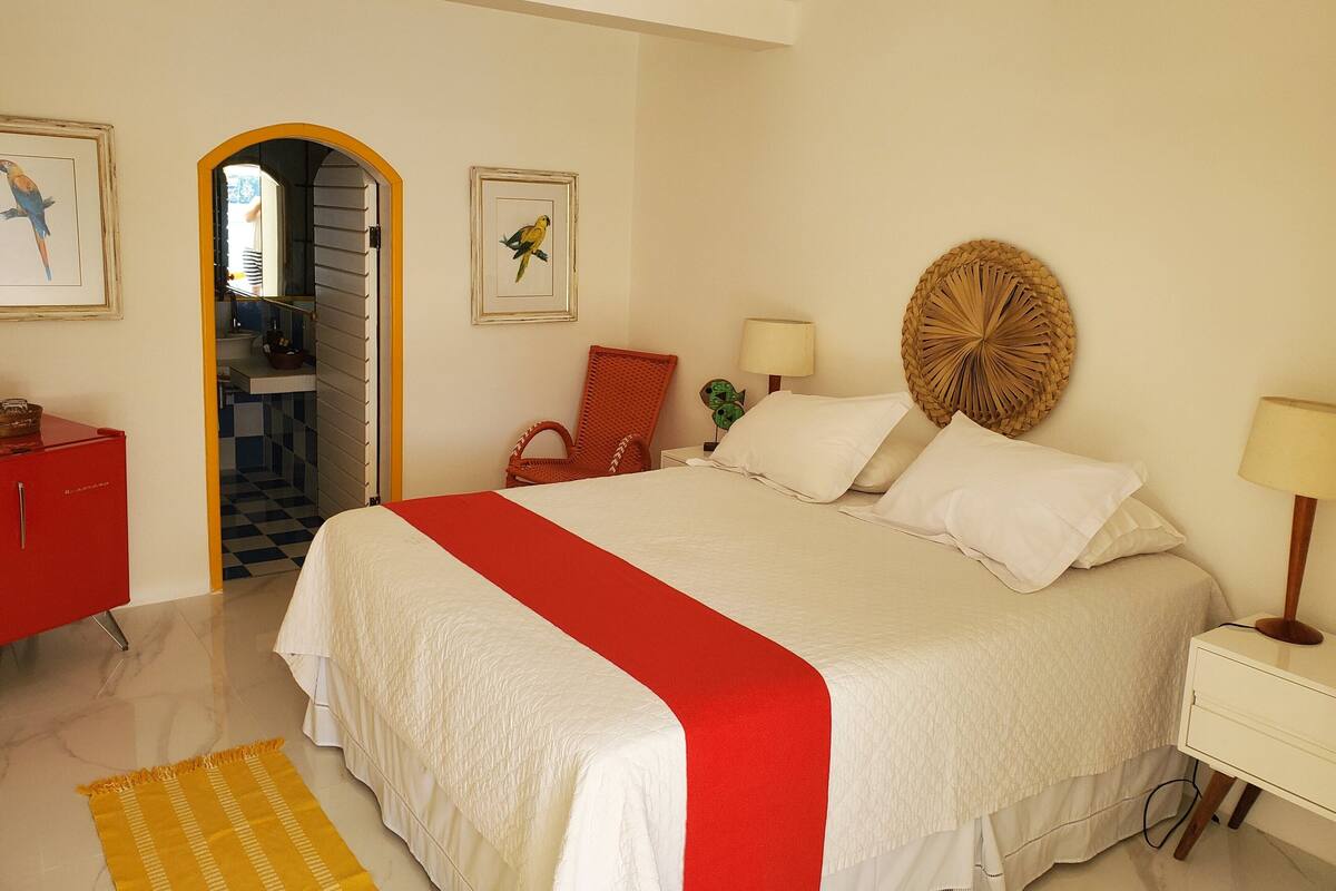 Un dormitorio con una cama grande, una colcha roja y blanca, un adorno de pared tejido, una mesita de noche con una lámpara y un armario rojo.