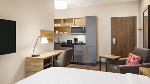 Habitación de hotel compacta con kitchenette, una cama, un escritorio con lámpara y una zona de estar con sofá y cojín.