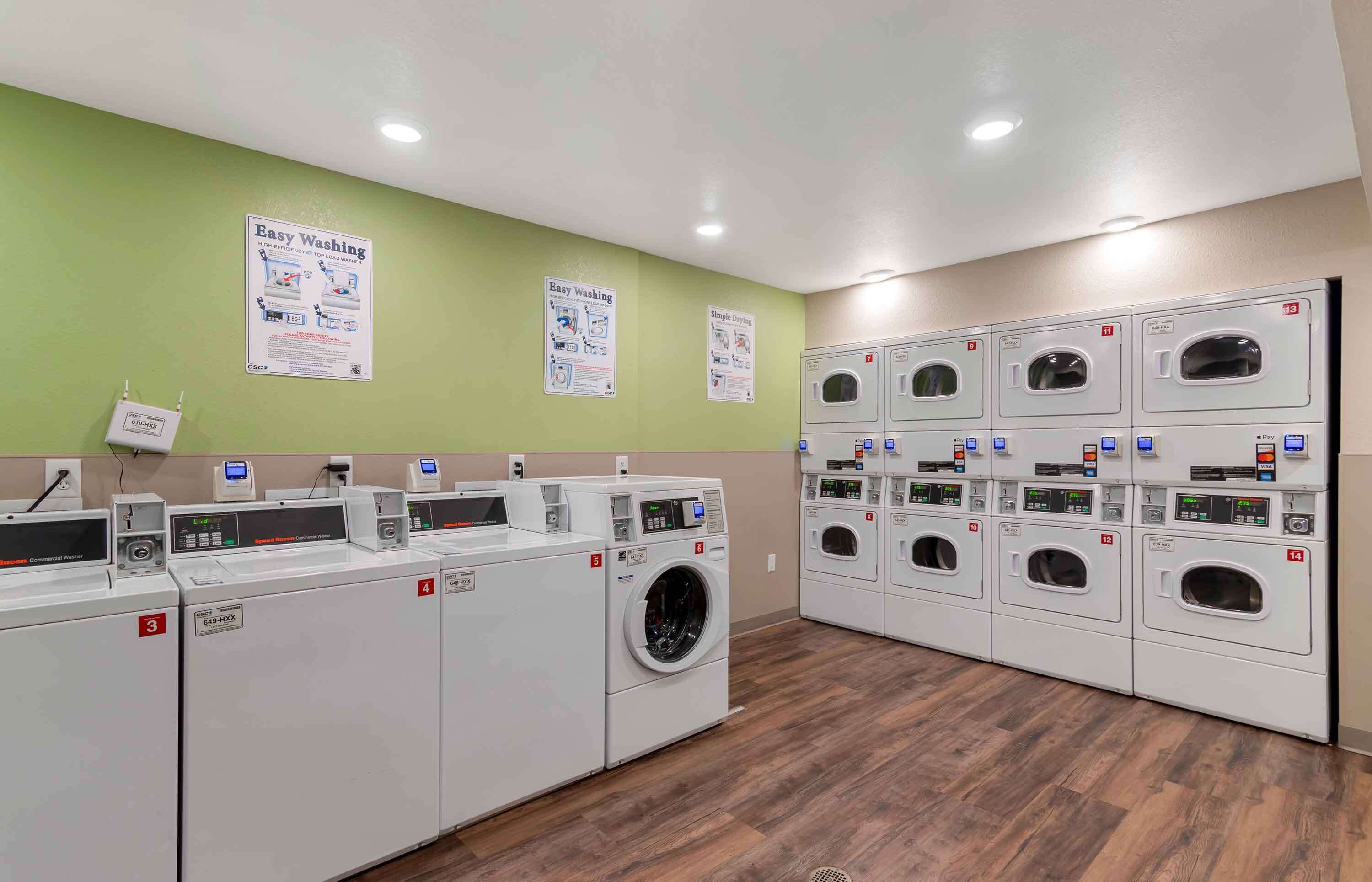 Een wasserette met meerdere wasmachines en drogers, genummerd van 3 tot 14, en instructieposters aan de muur.