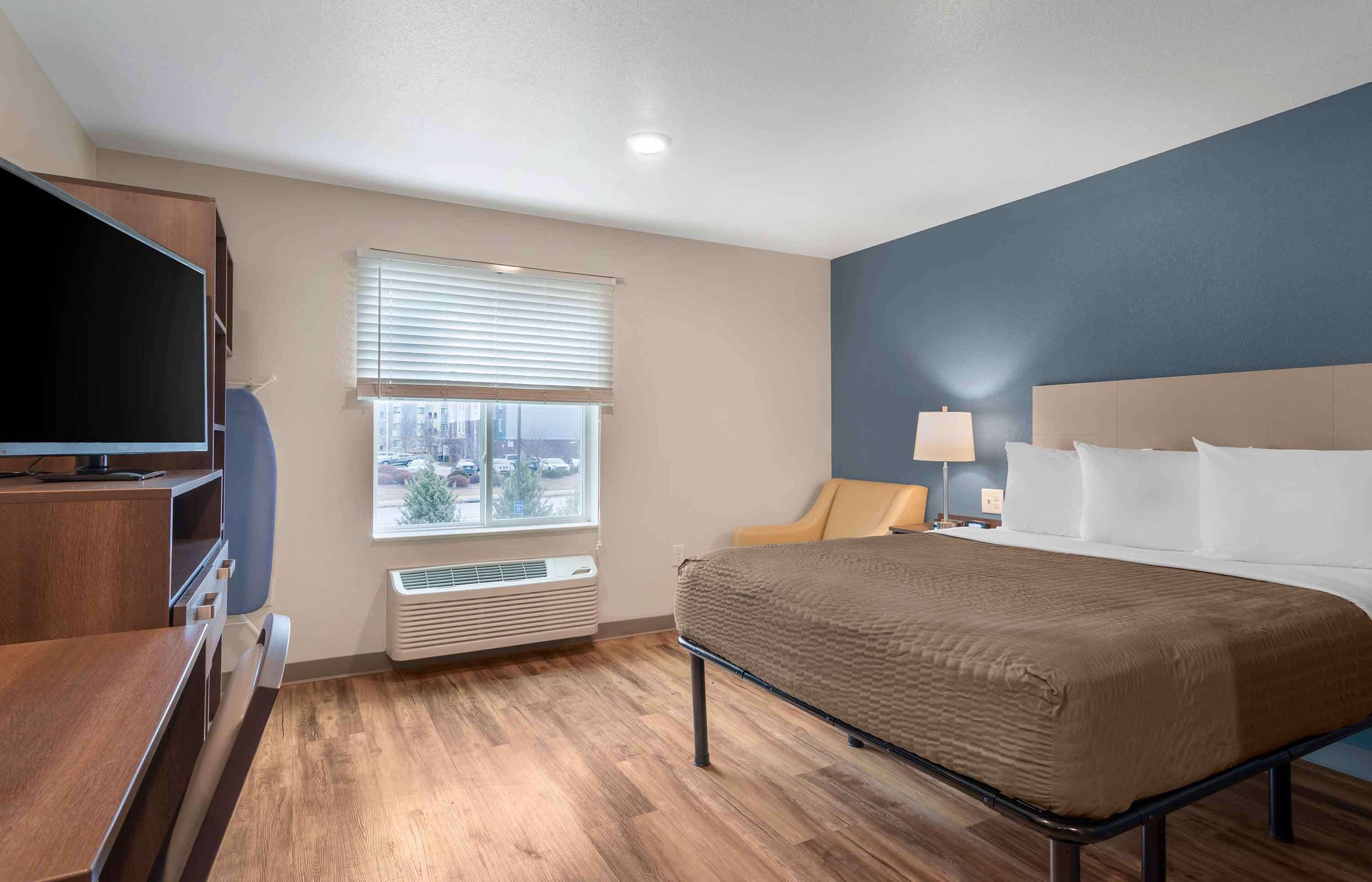 A hotel room with a bed, a desk with a TV, a window with blinds, and a lamp on the nightstand.