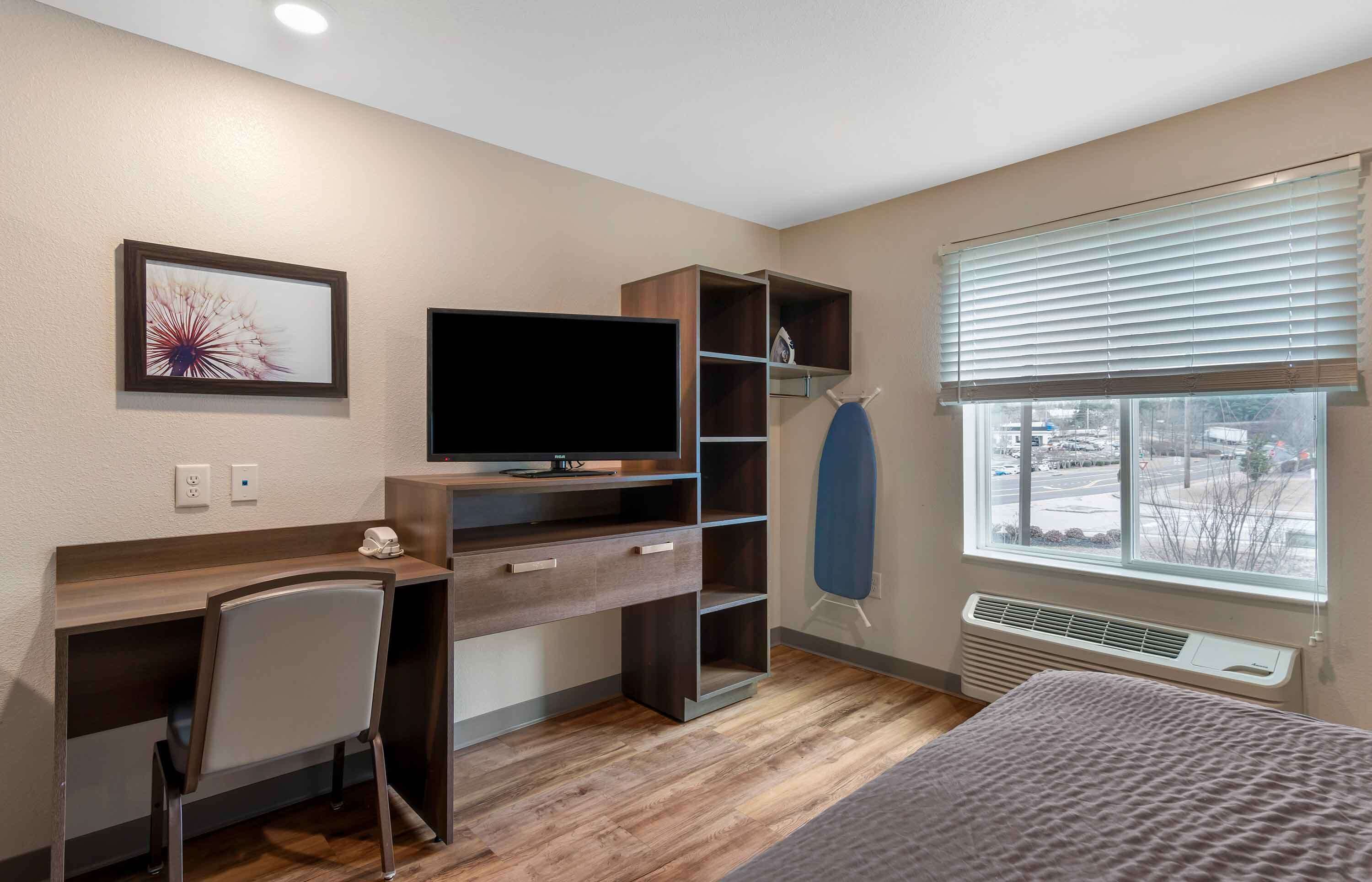 A bedroom with a wooden desk, a chair, a television, a wardrobe, and a window with blinds.