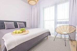 A modern bedroom with a bed, a bedside table with fruit, a wicker chair, and sheer curtains. - Novis Apartments Panorama View (Warsaw)