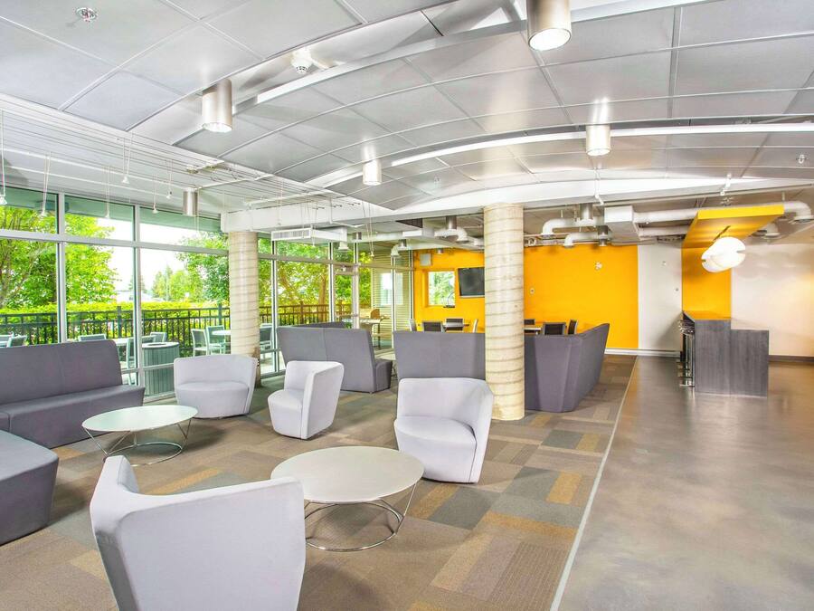 Un espace intérieur moderne avec des canapés gris, des fauteuils blancs et une table basse centrale. De grandes fenêtres laissent entrer la lumière naturelle et offrent une vue sur la verdure environnante.