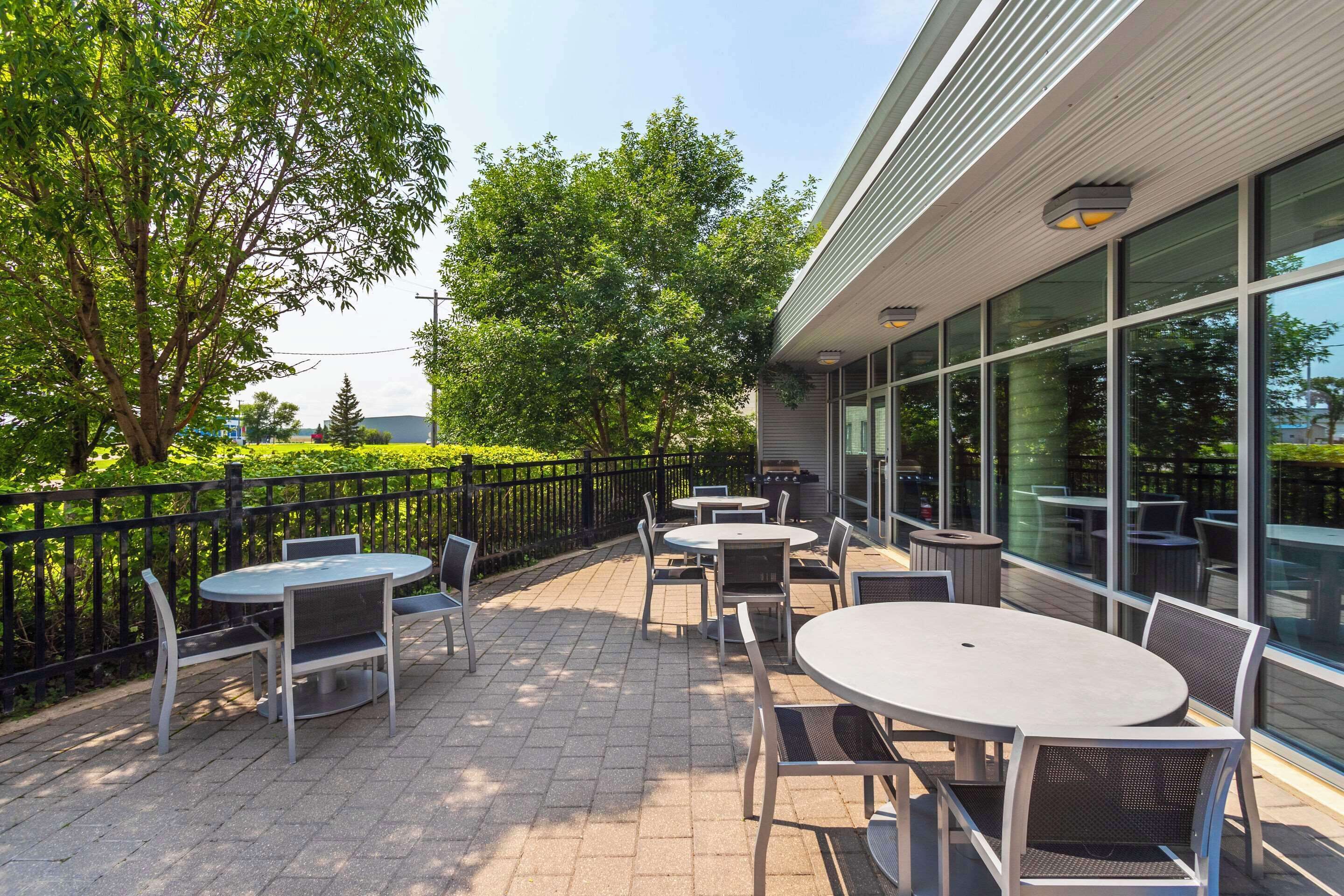 An outdoor patio with round tables and chairs, surrounded by a black metal fence, and large windows on the building.