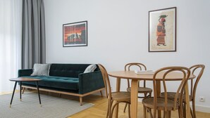 A living room with a dark green sofa, a small wooden table, and four wooden chairs. There are two framed pictures on the wall. - Novis Apartments Przy Rotundzie (Warsaw)