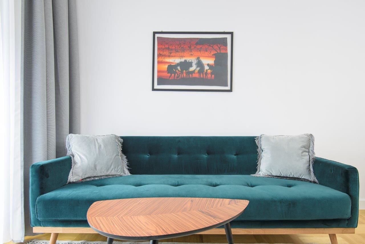 A teal sofa with two pillows, a wooden coffee table, and a framed picture of a sunset scene on the wall.