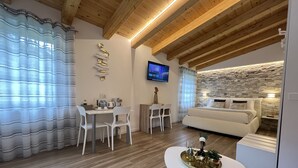 A modern living room with a dining area, wooden ceiling beams, a stone accent wall, and a flat-screen TV. - The Garden (Riomaggiore)