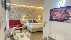 A modern living room with a stone wall, a red sofa, a white bed, and a dining area with a table and chairs. - The Garden (Riomaggiore)