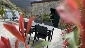 A stone building with a green shutter, a patio with a table and chairs, and a mountainous backdrop. - The Garden (Riomaggiore)