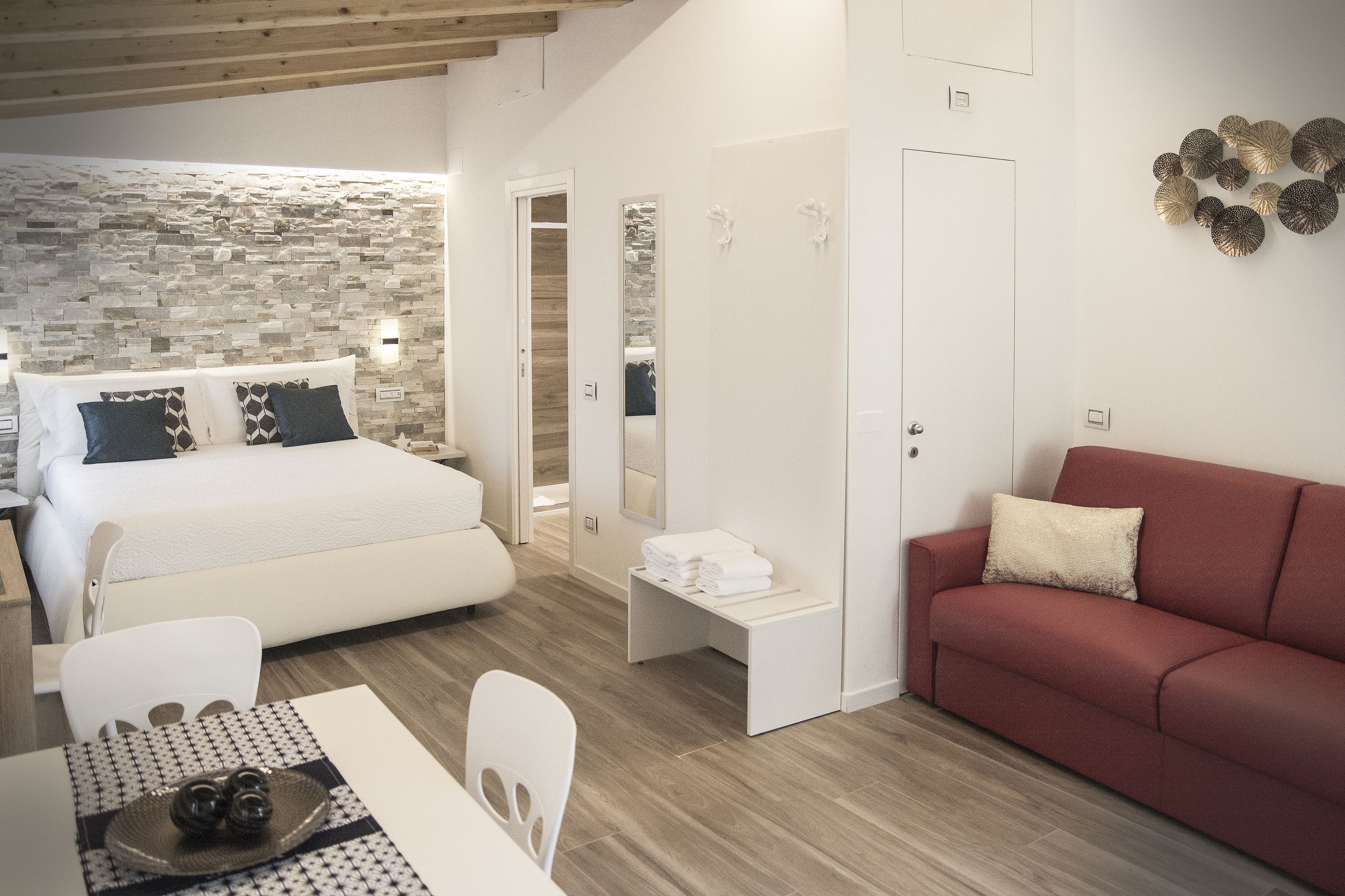 A modern hotel room with a stone wall, a bed with patterned pillows, a red sofa, a small table with a tray, and a mirror.