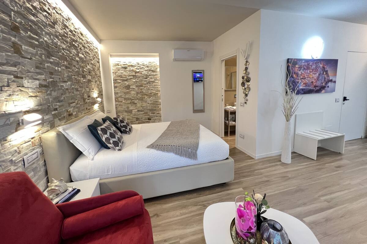 Un dormitorio moderno con una pared de piedra, una cama con almohadones estampados, una silla roja y una mesita de noche blanca con un florero.