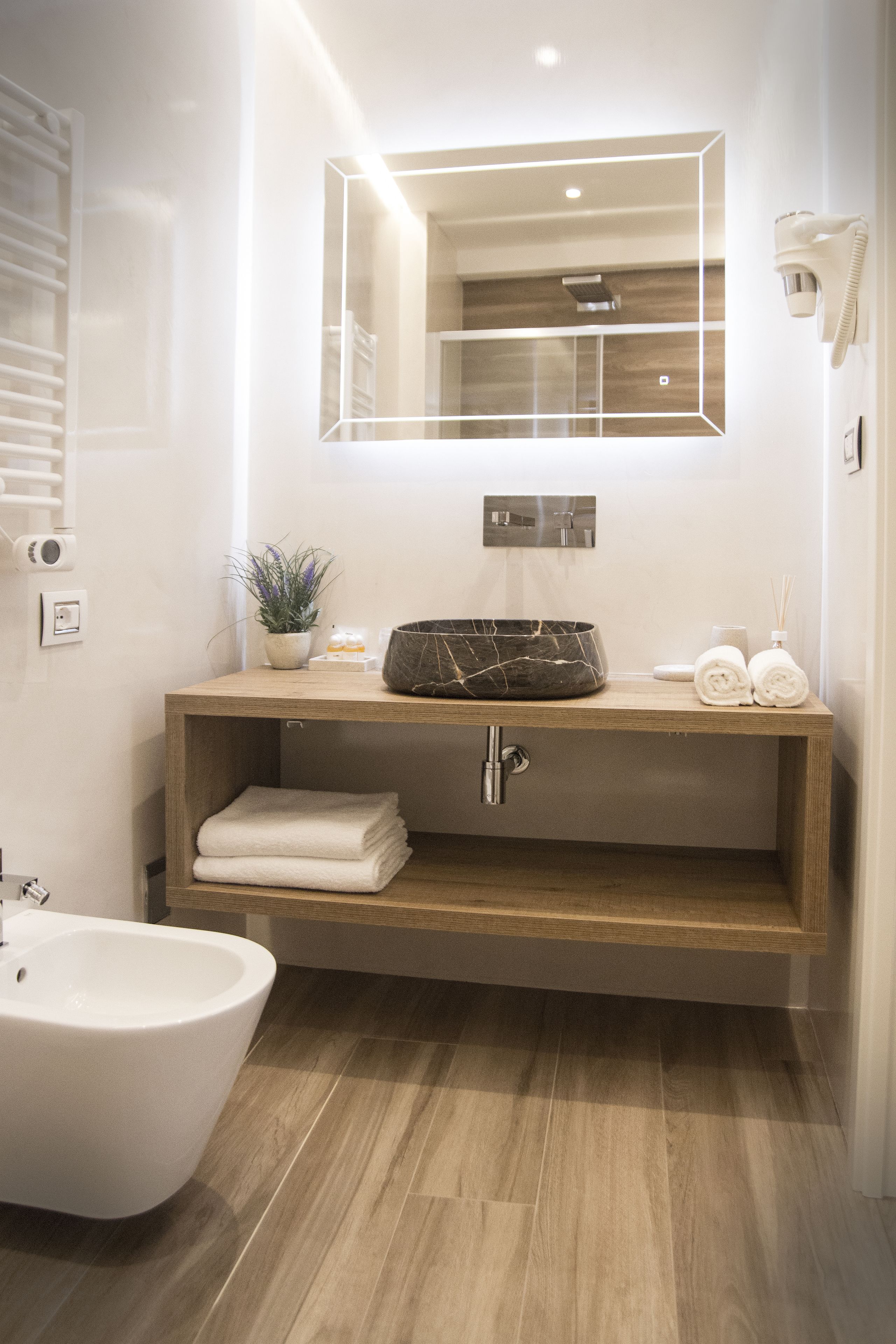 Un baño moderno con tocador de madera, lavabo de mármol negro, un bidé blanco e iluminación integrada en el espejo.