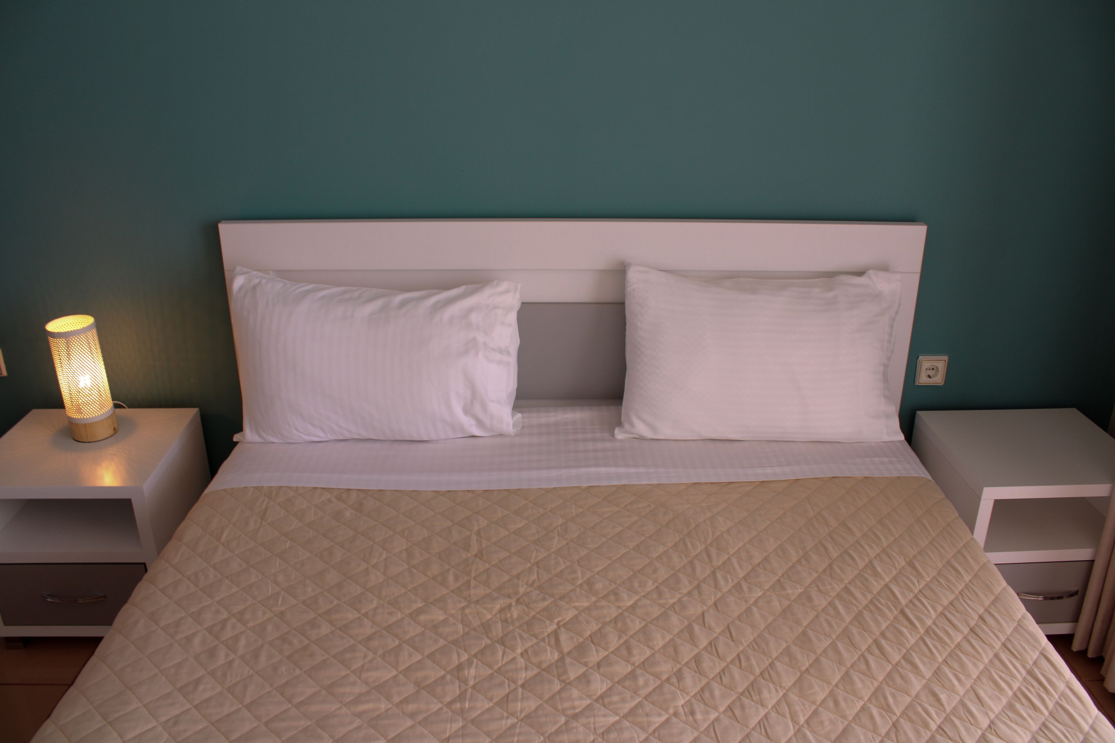A bed with a quilted beige cover, two white pillows, and a white headboard. There is a bedside lamp on the left side of the bed.