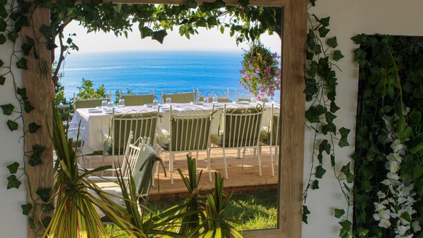 Un espace repas offrant une vue sur la mer, entouré de verdure et agrémenté de mobilier blanc.