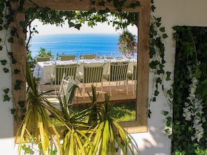 Un espace repas offrant une vue sur la mer, entouré de verdure et agrémenté de mobilier blanc.