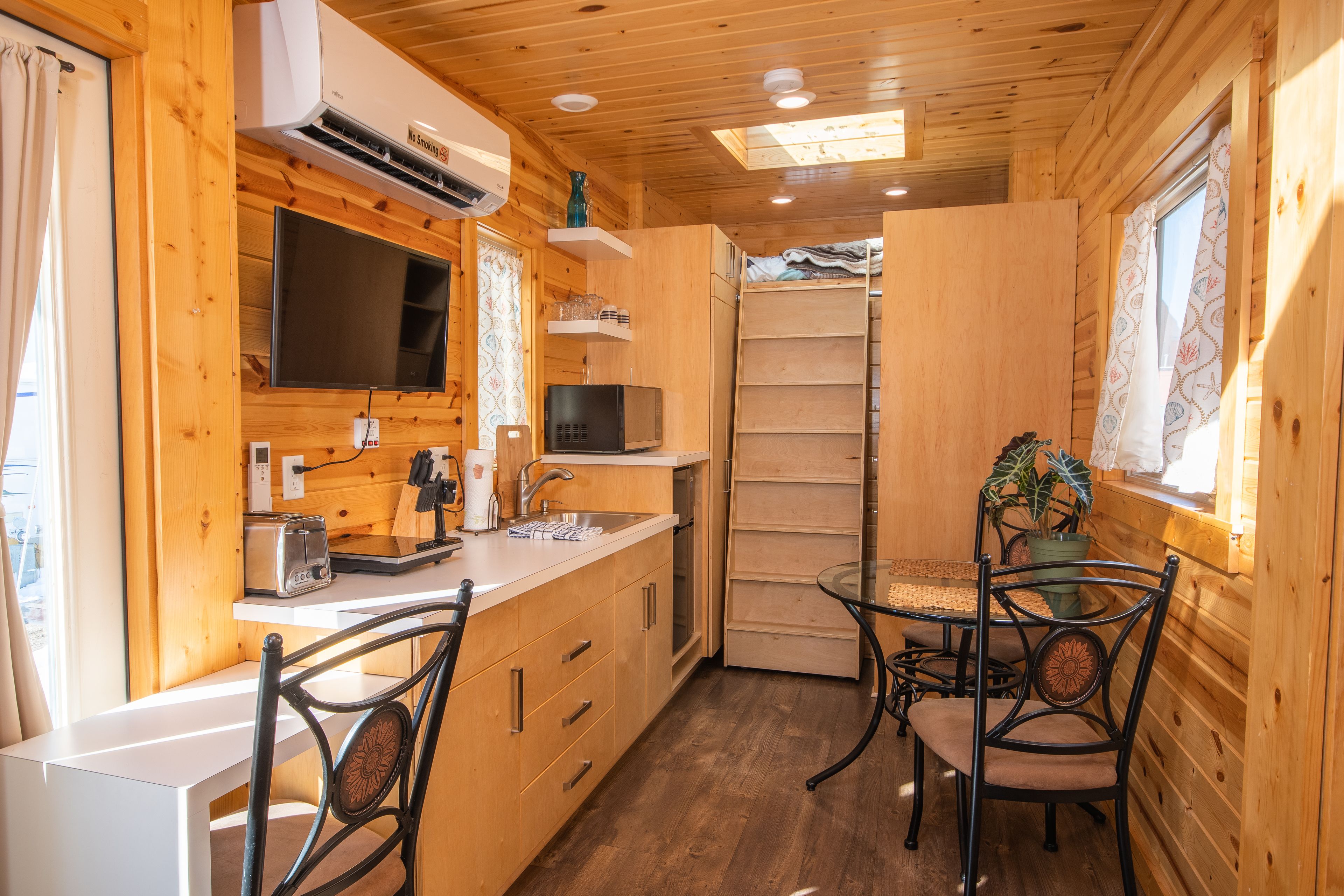 Una cabaña de madera compacta con kitchenette, un comedor con mesa y sillas de vidrio, y un pequeño espacio de sala con televisor y estanterías.
