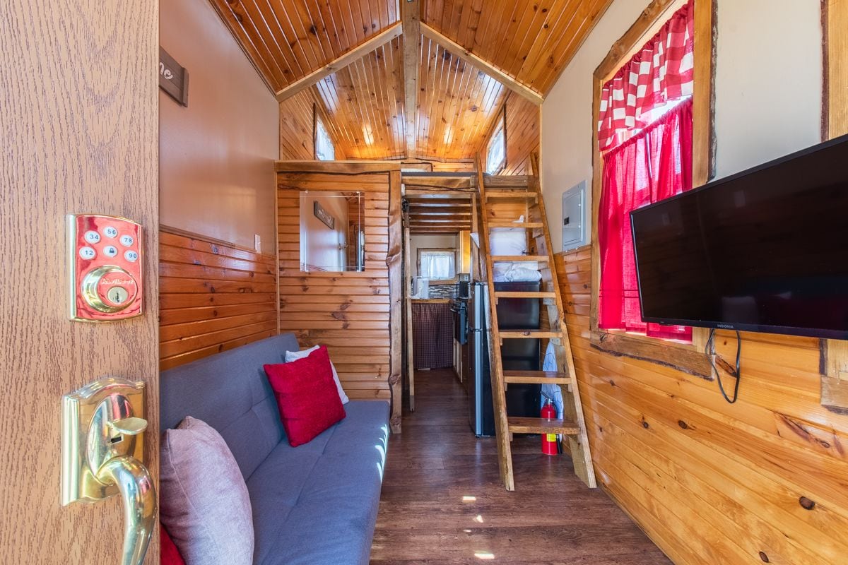 Un accogliente interno di una baita con soffitto in legno, un divano con cuscini rossi, una televisione e una scala che conduce a una mezzanine.