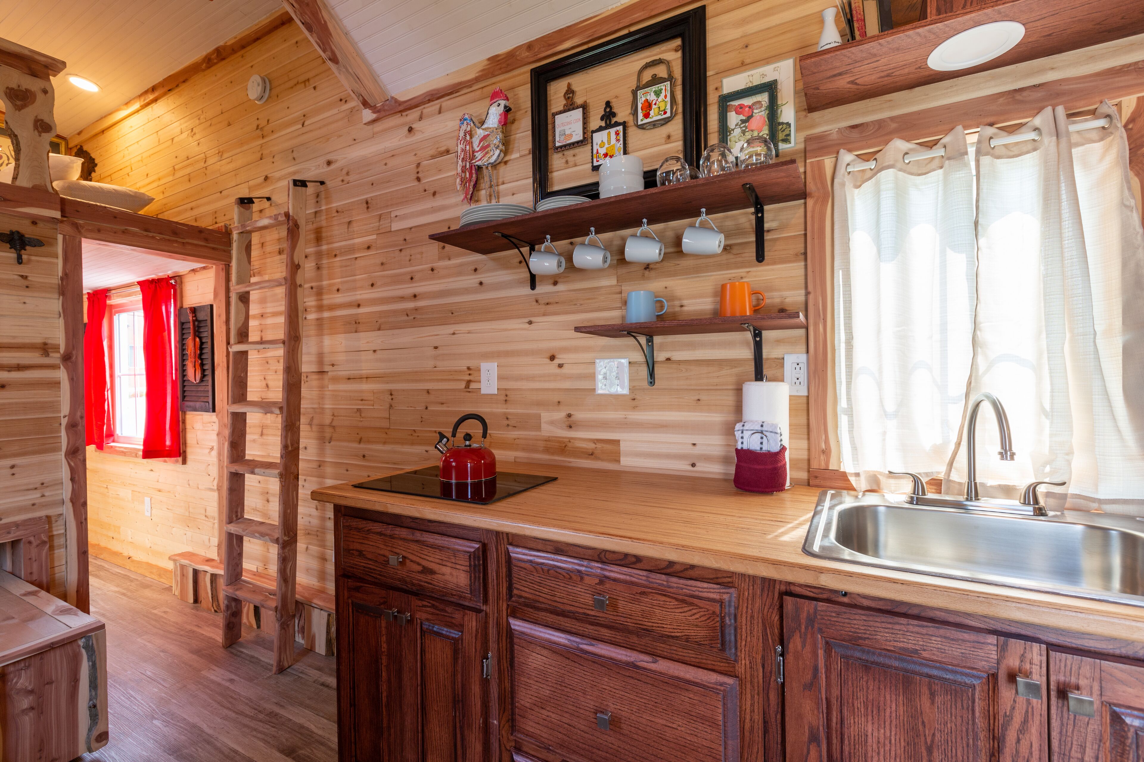 A wooden interior with a kitchen area featuring a sink, a red kettle, and a shelf with cups.