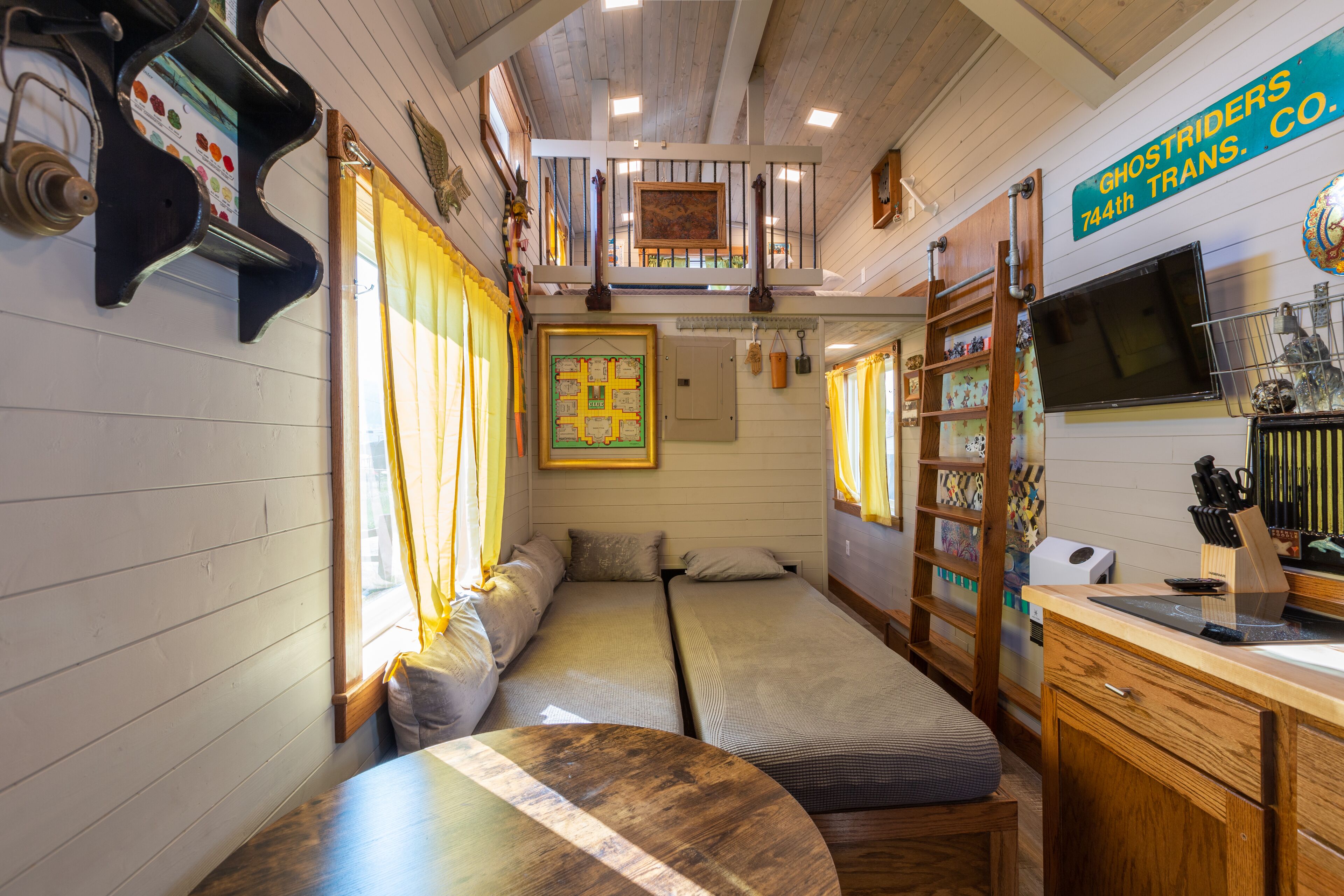 Acogedor interior de cabaña con dos camas individuales, una mesa de comedor de madera, una zona de cocina con fregadero y estufa, y un altillo sobre las camas.