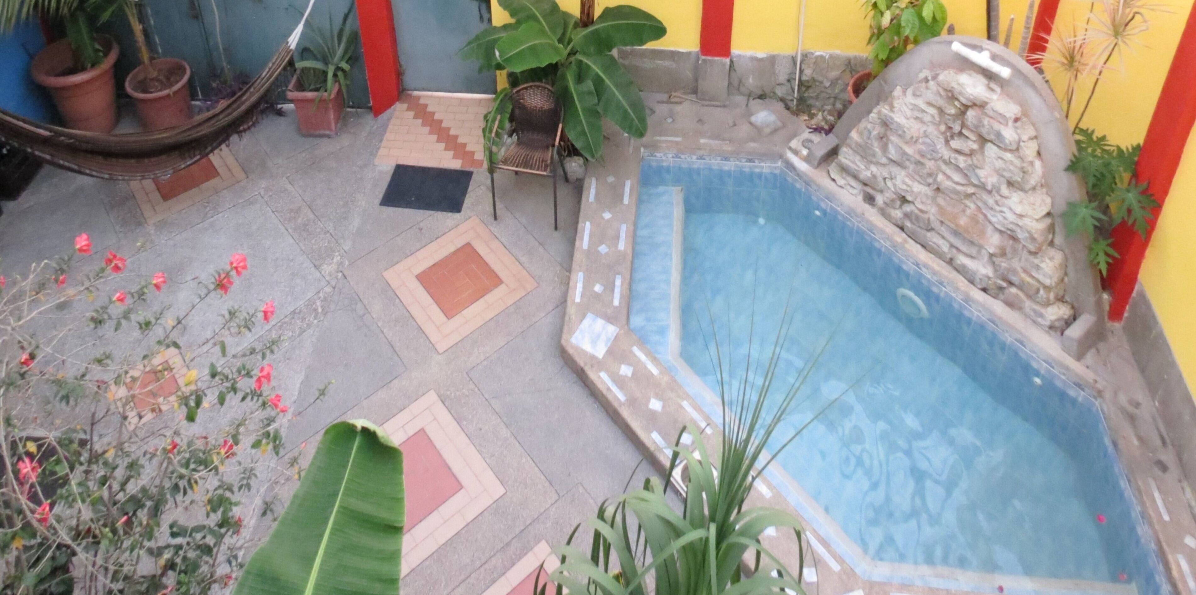 Una piscina pequeña con una pared de piedra y una zona de terraza con baldosas, plantas en macetas y una silla.