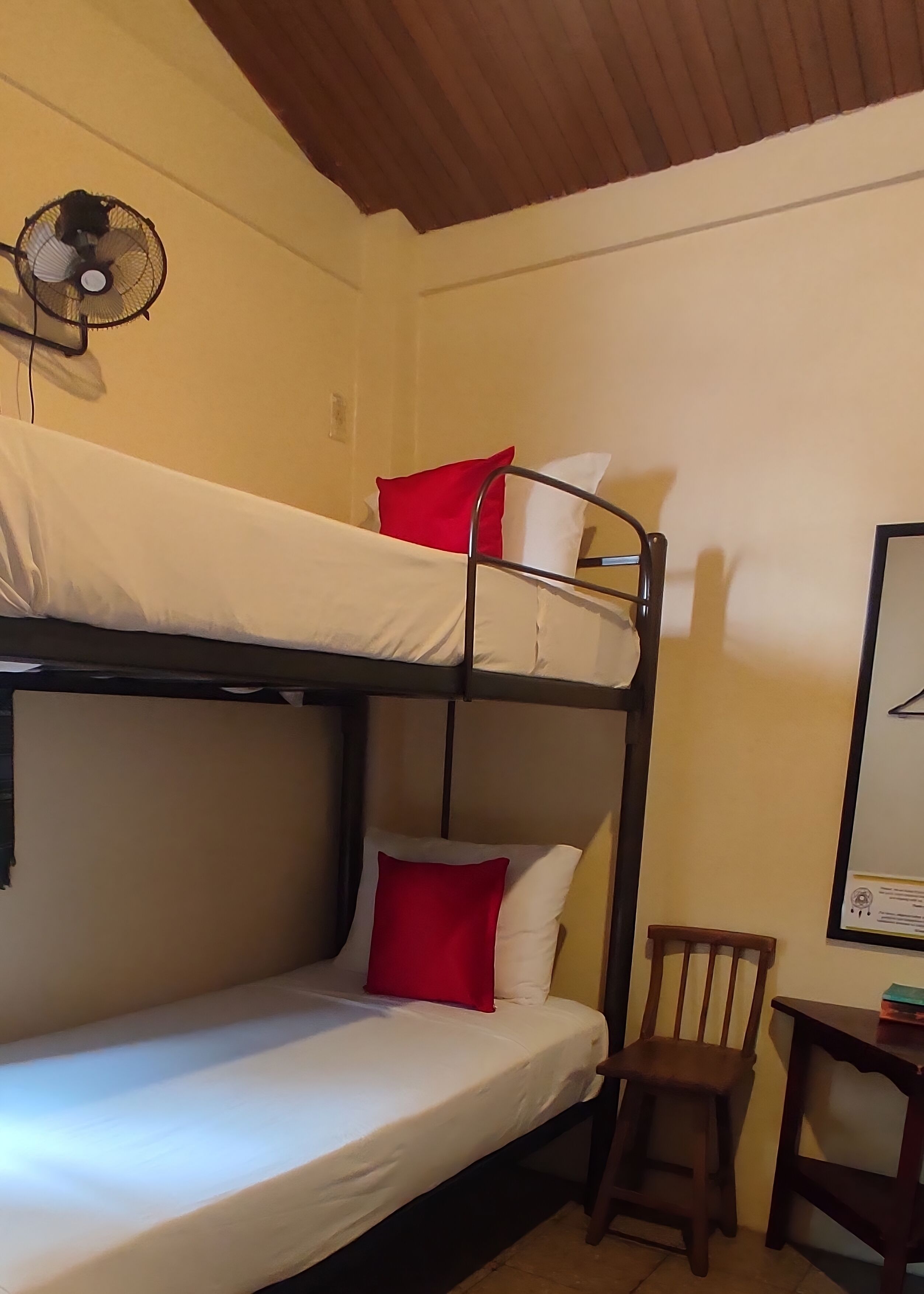 Habitación con literas, un ventilador, una silla de madera y una mesita pequeña.