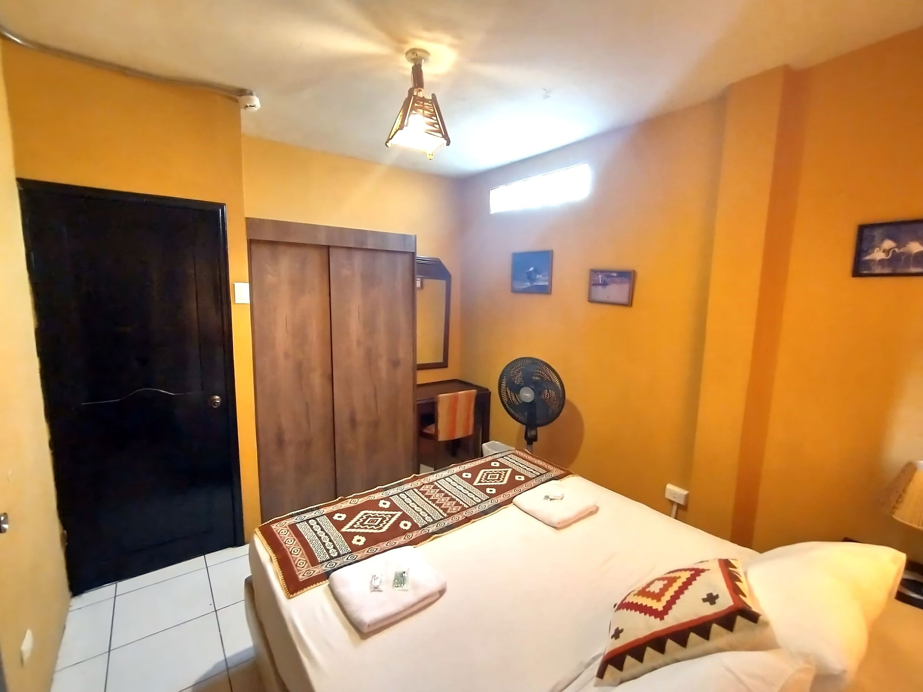 A hotel room with a bed, a fan, a window, and a door.
