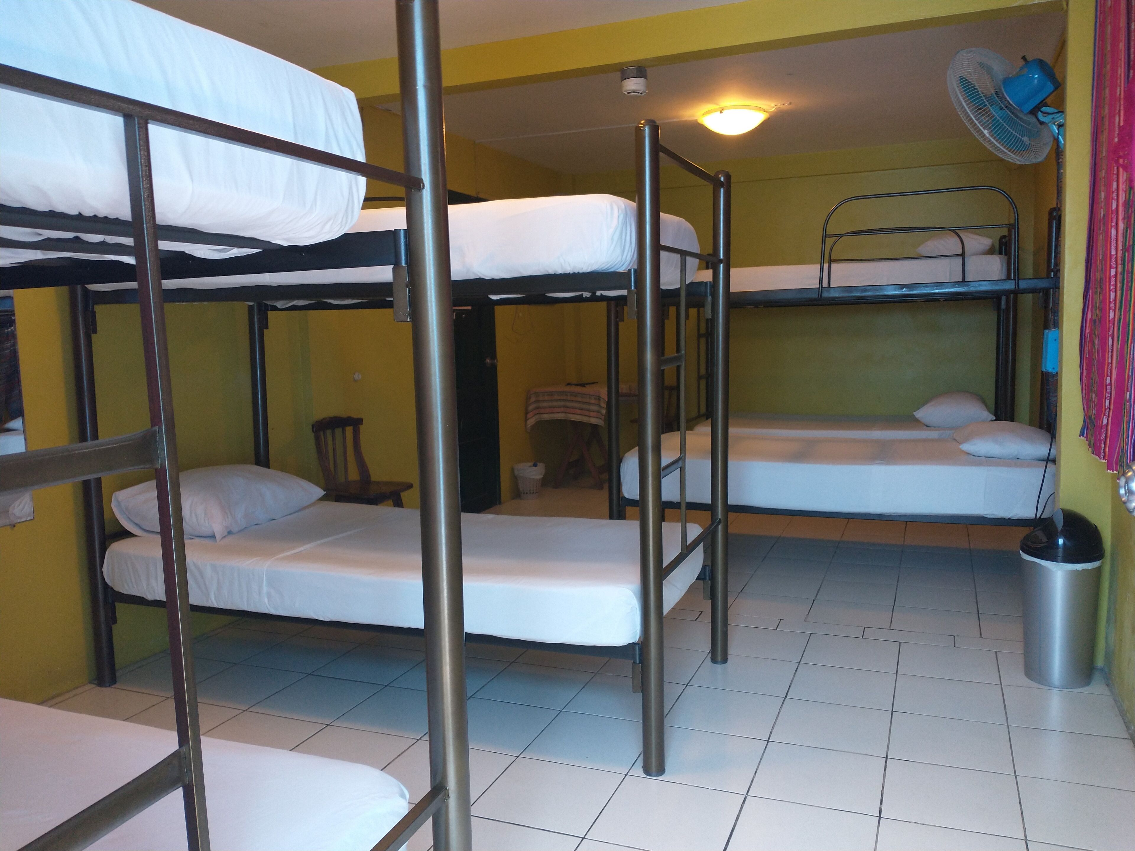 Una habitación tipo dormitorio con literas, un ventilador, un cubo de basura y una silla.