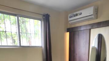 Un dormitorio con cama, ventana con cortinas, unidad de aire acondicionado, espejo y toma de corriente.