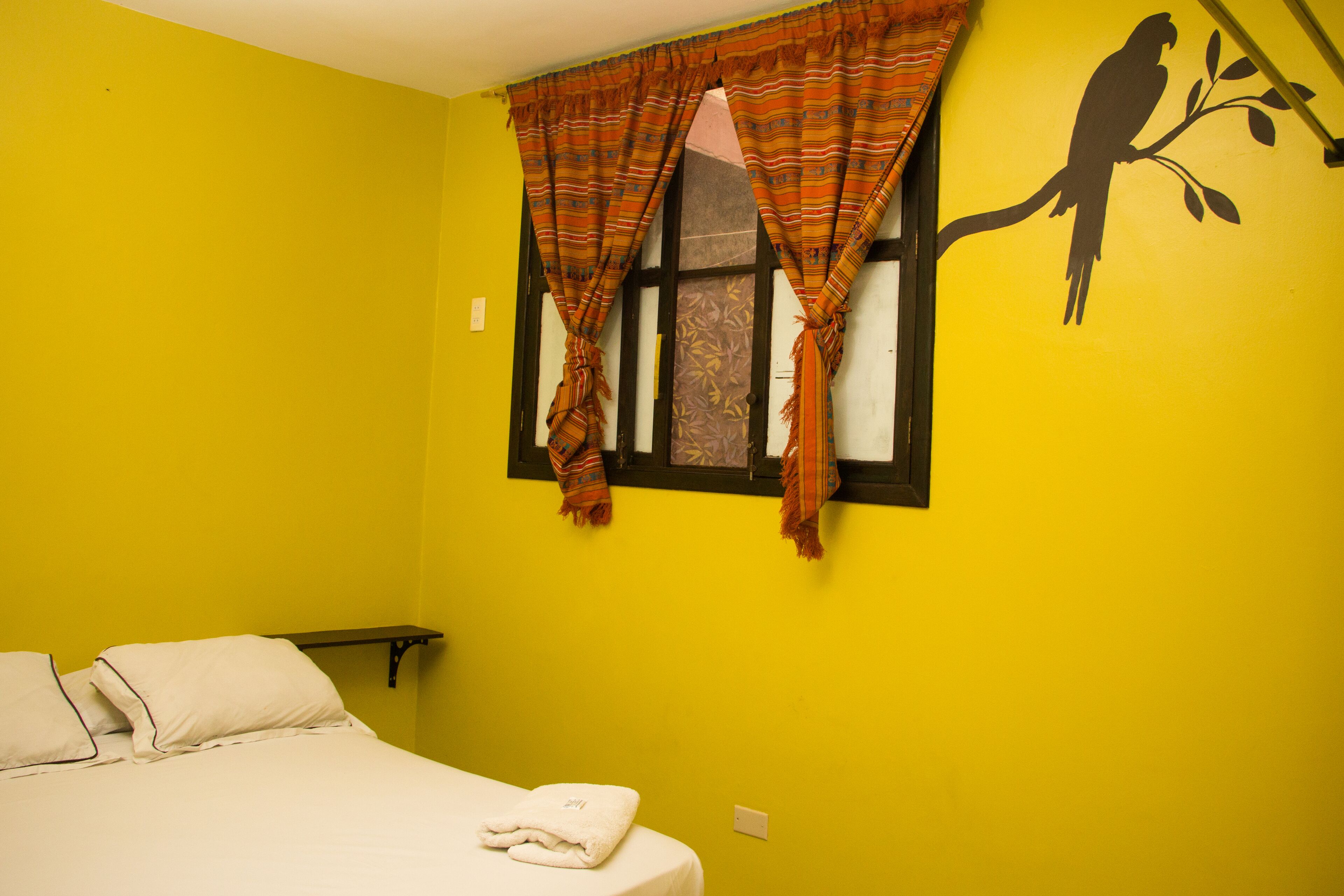 Una habitación con cama, ventana con cortinas y un adorno de pared con un pájaro en una rama.