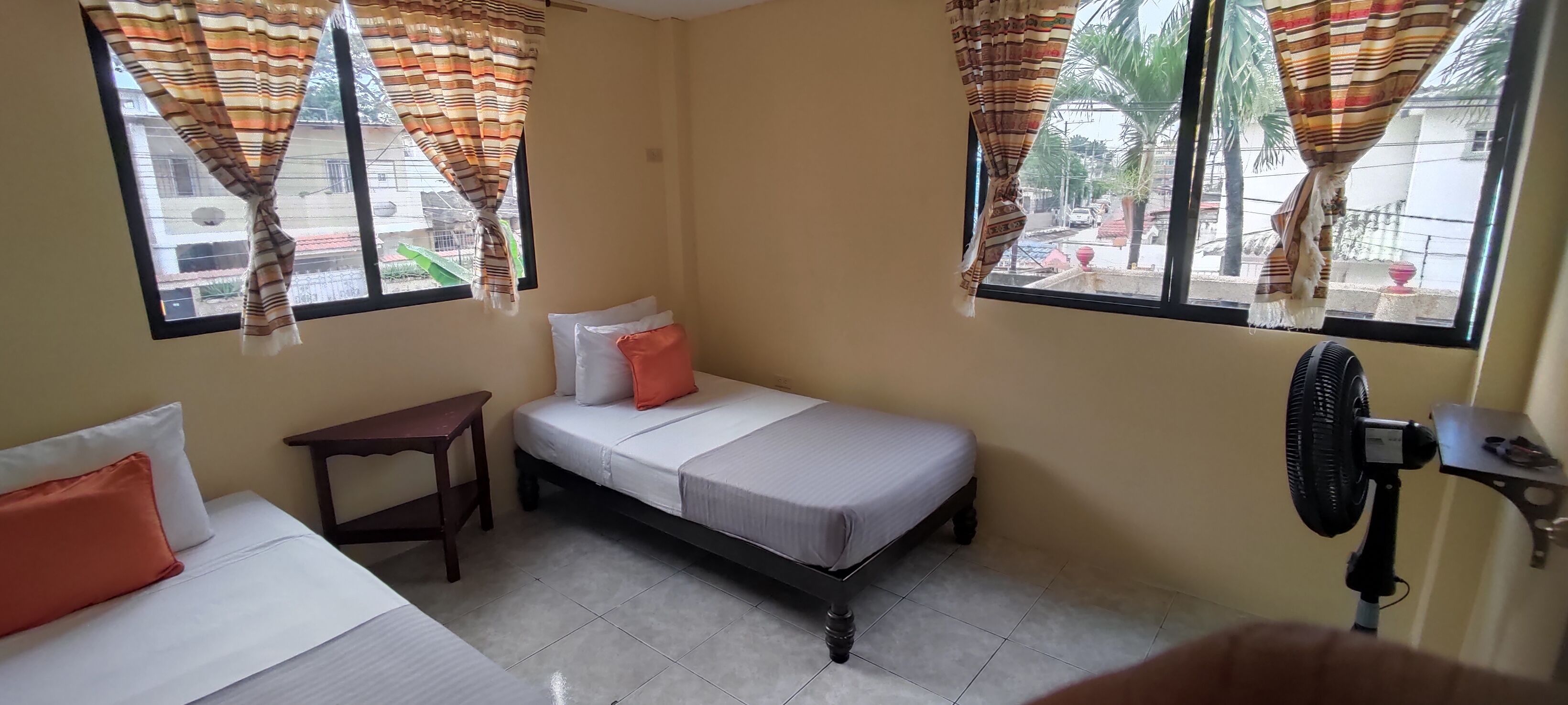 A small, single-room accommodation with a bed, a bedside table, and a standing fan.