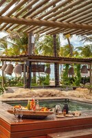 A wooden deck with a dining setup overlooking a pool, surrounded by palm trees and a beach view.
