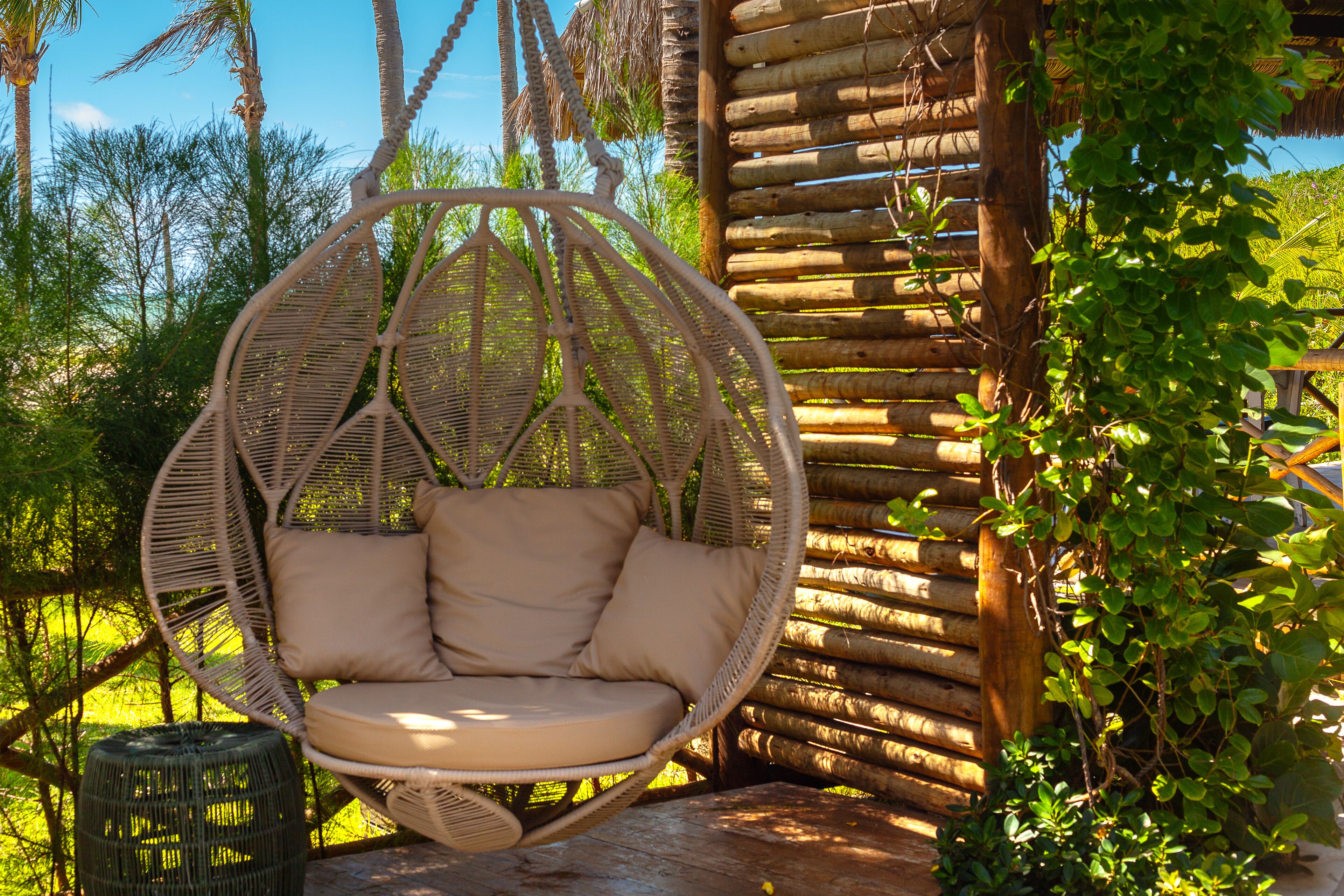 A wicker hanging chair with cushions, a wooden fence, and lush greenery in the background.