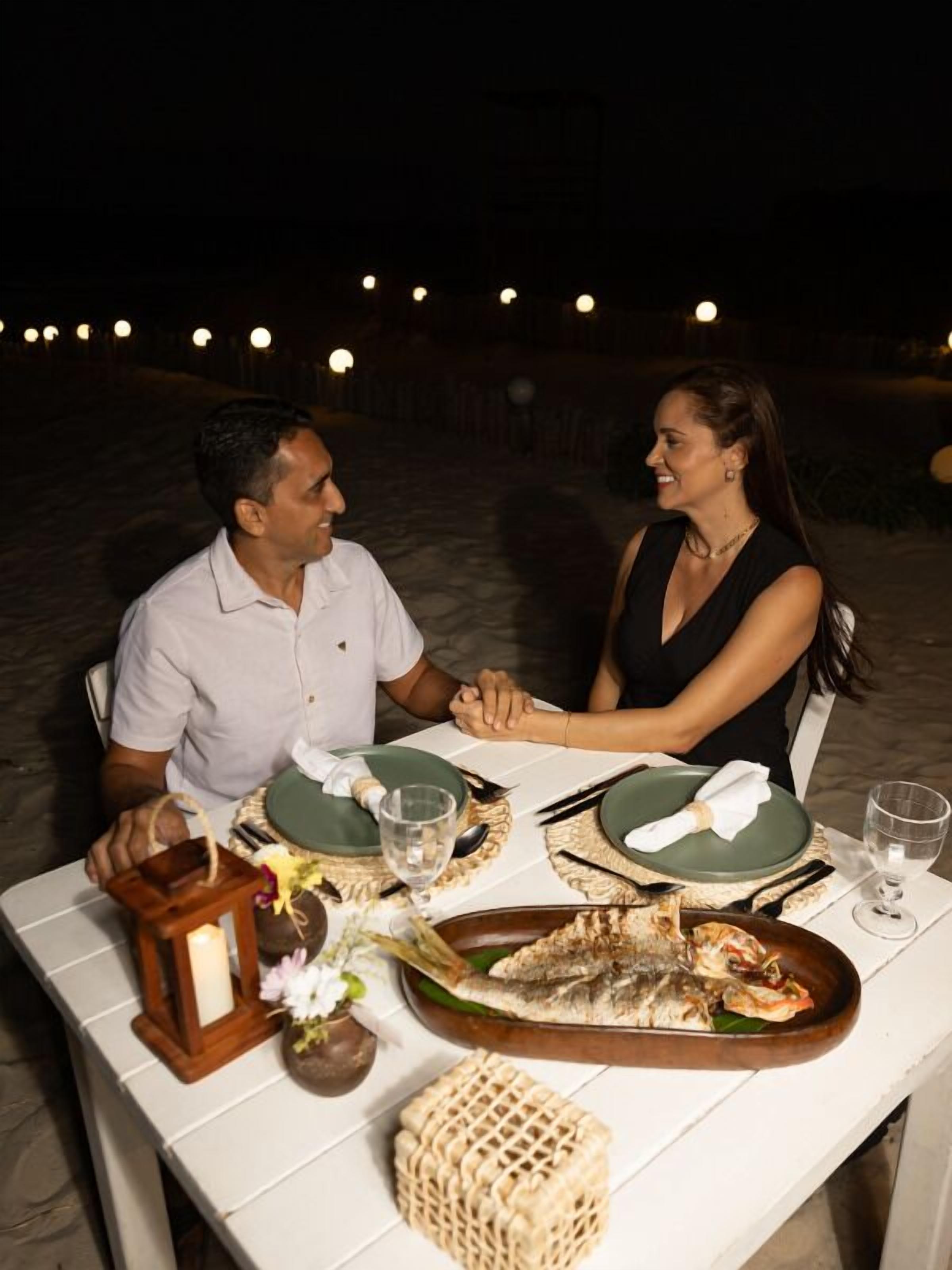 Un couple est assis à une table en plein air, devant un plat de fruits de mer, et l’ensemble est illuminé par des guirlandes lumineuses, avec une plage en arrière-plan.