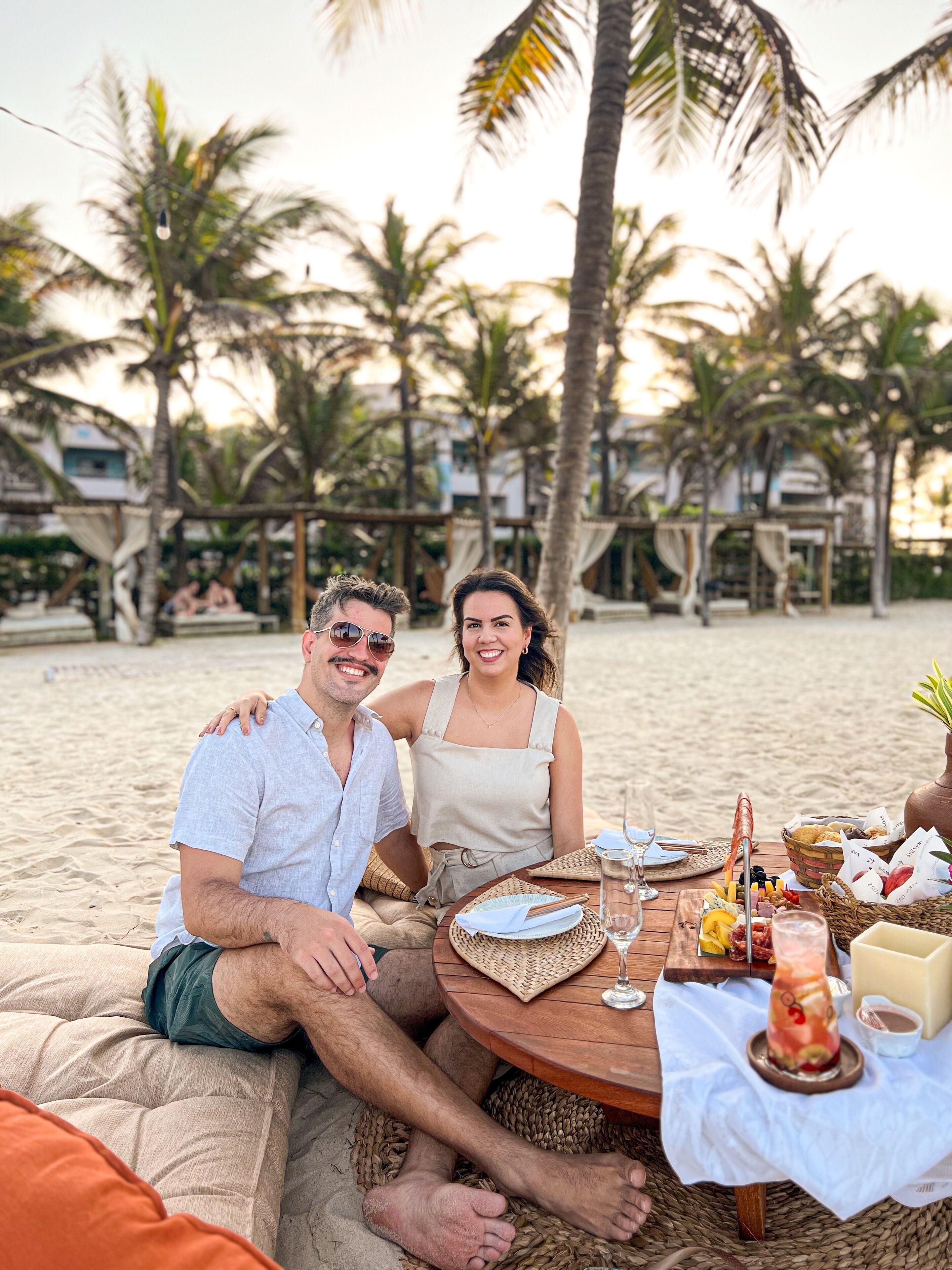 A couple sitting at a wooden table on a sandy beach with palm trees in the background.